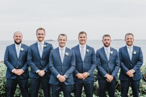 Groomsmen in matching navy suits pose for formal portrait by the ocean.