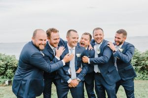 Groomsmen in navy suits share a laugh during candid moment at seaside wedding.
