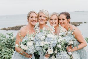 Bridal party in light blue dresses hold matching bouquets against coastal backdrop.