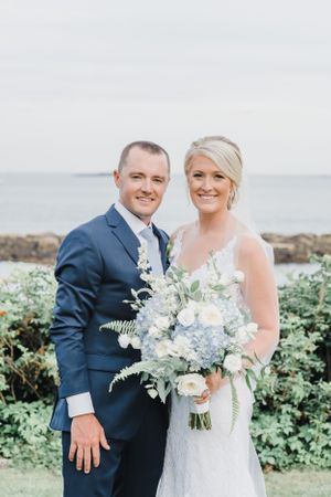 Newlyweds pose for portrait with blue and white floral bouquet against coastal background.
