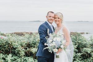 Wedding portrait of couple in navy suit and white gown holding pastel bouquet by the ocean.