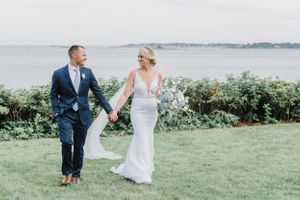 Wedding couple walks hand in hand on coastal lawn with ocean view in background.