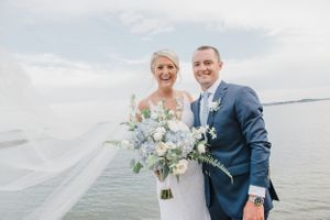 Wedding couple poses by the waterfront in formal attire with a soft blue and white bouquet.