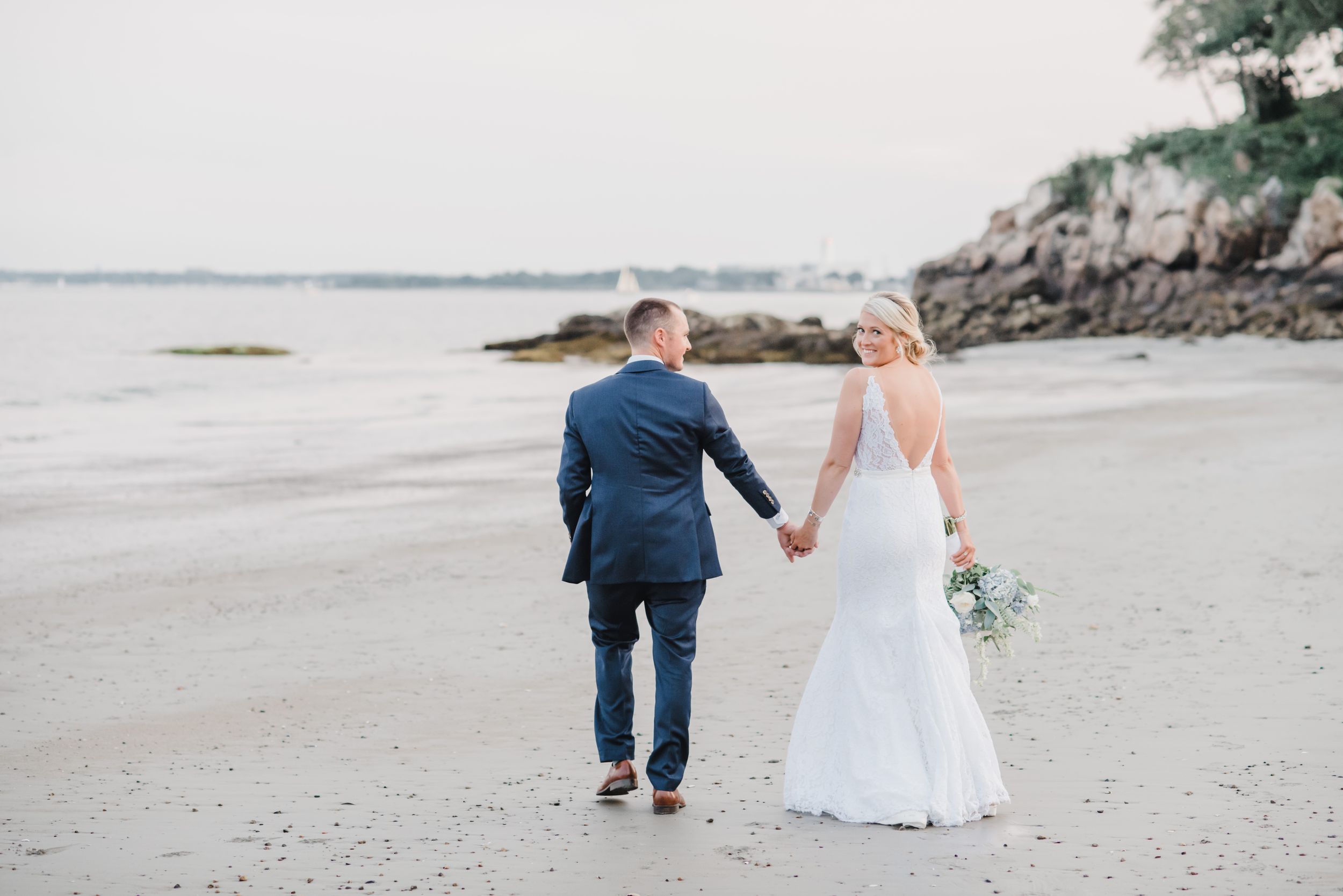 Newlyweds walking hand in hand along a scenic beach shoreline at sunset.