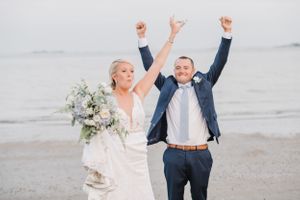 Joyful wedding couple celebrating with raised arms on a beach while holding a white and blue floral bouquet.