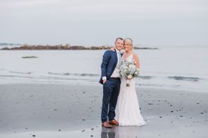 Couple standing together on misty beach with ocean rocks in background.