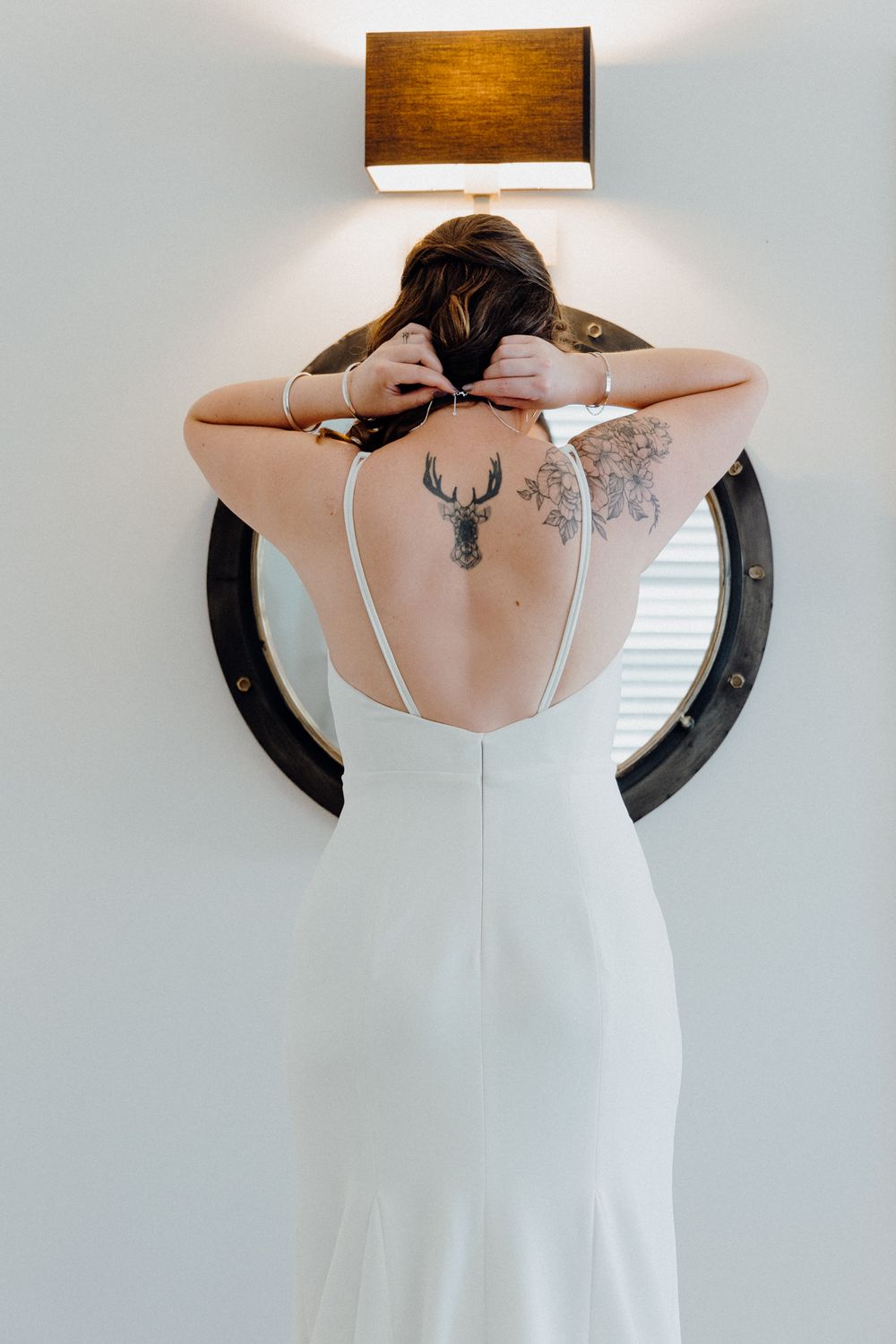 Bride and her deer and floral tattoos, with round mirror and wall lamp above, captured by Apertured Media Auckland Wedding Photographer.