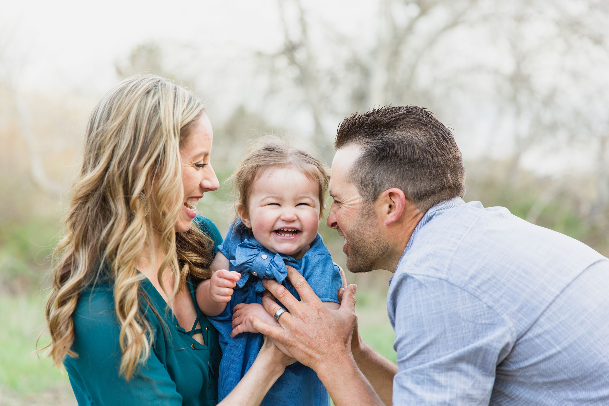 A joyful family moment outdoors with parents sharing a laugh with their toddler wearing blue against a nature backdrop for family pictures.