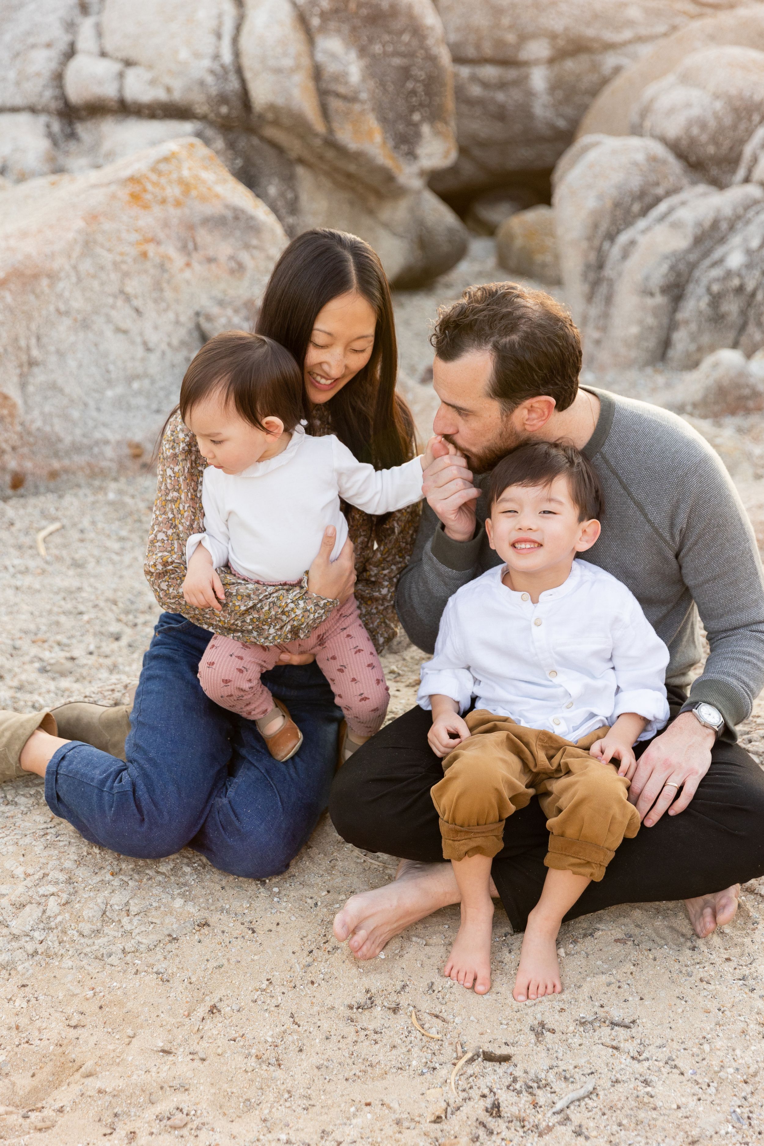 Family sitting together on sandy beach with large rocks in background during golden hour photoshoot in Carmel.