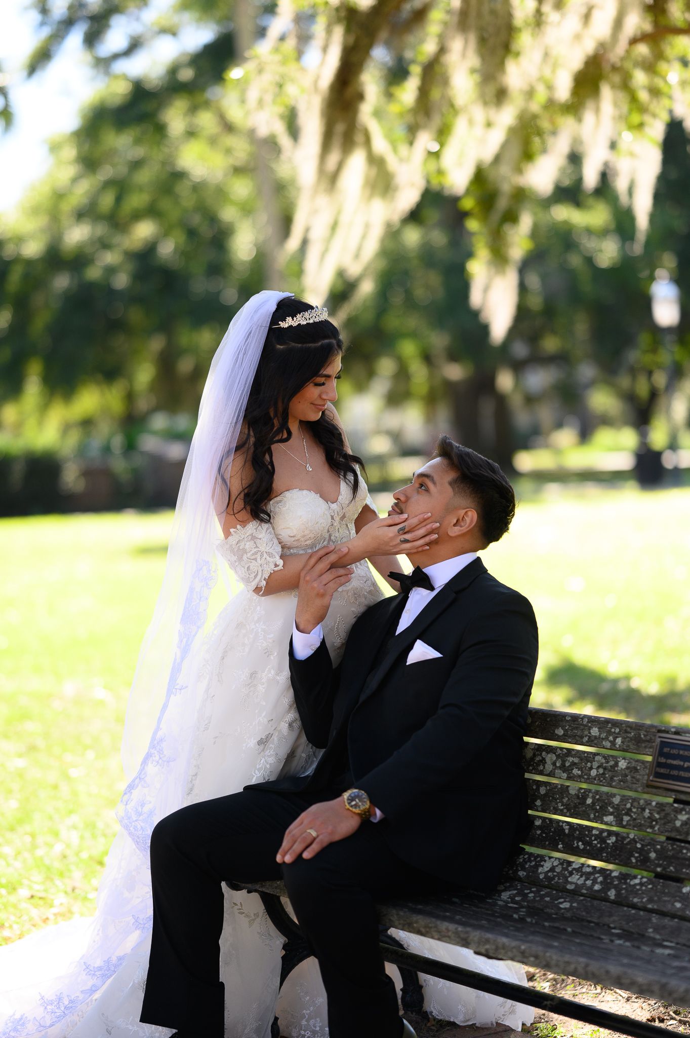 A romantic wedding photo of a couple on a park bench surrounded by hanging moss in a garden setting.