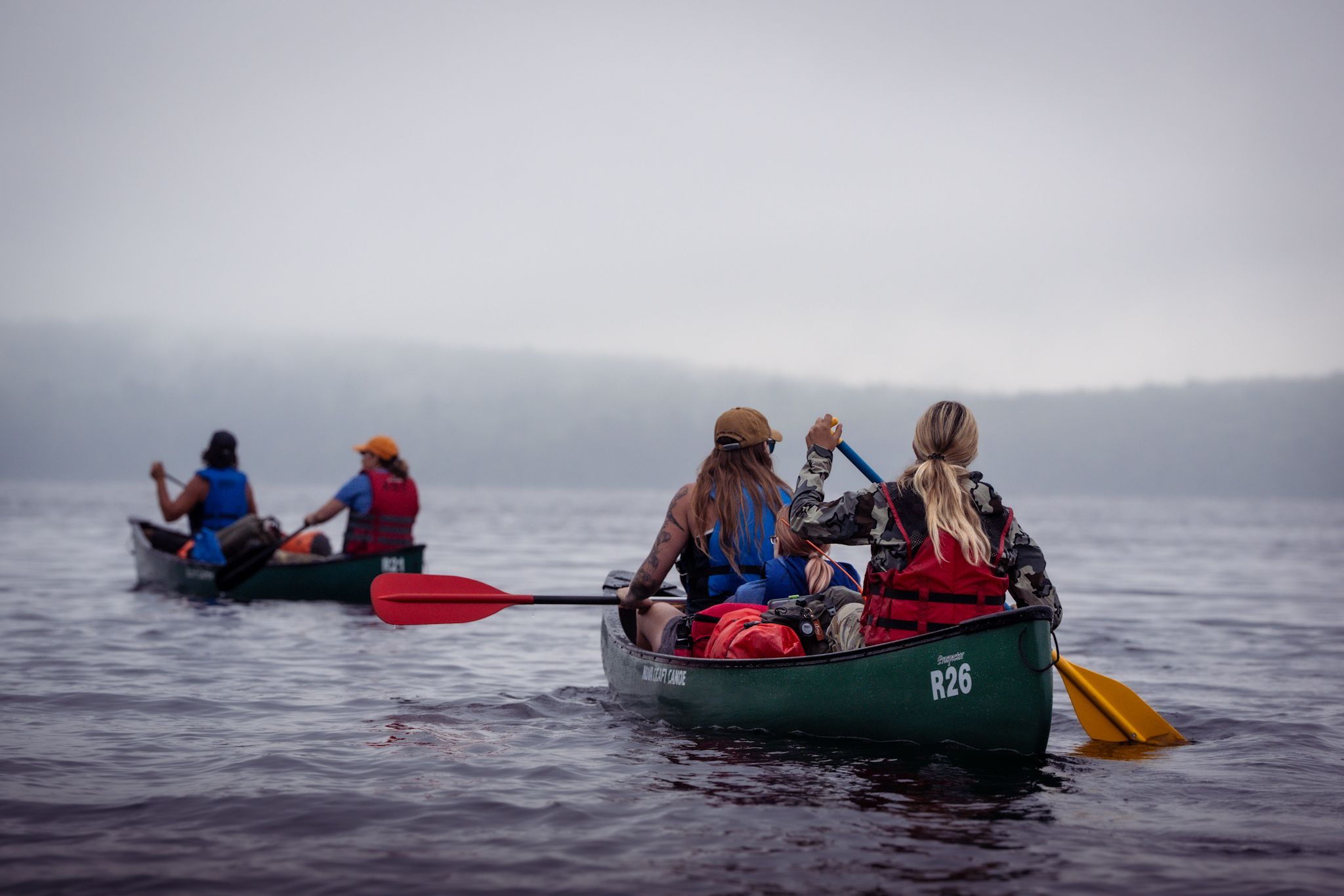 A group of people paddle canoes across a misty lake on a cloudy day with red and blue watercraft visible.