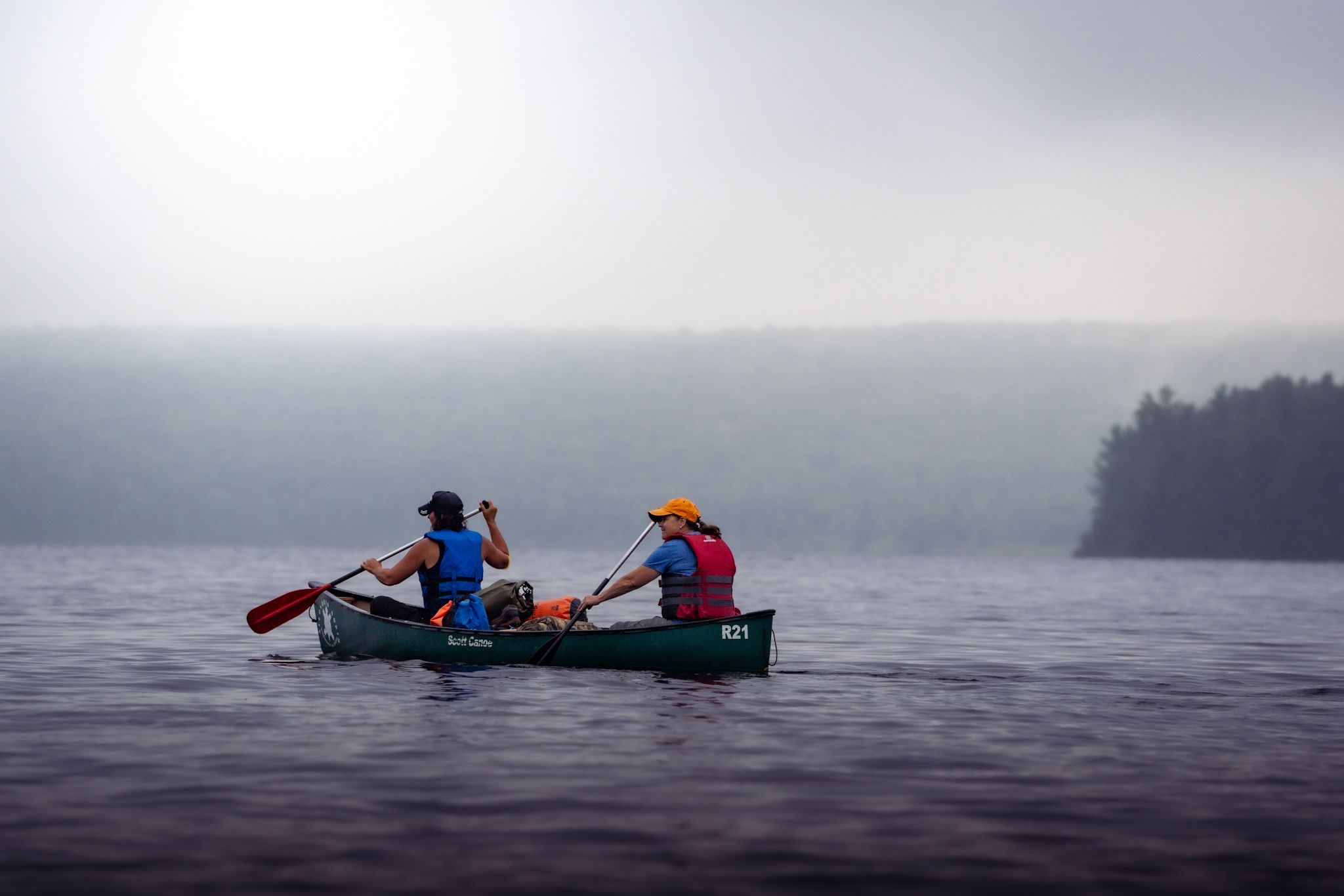 Two paddlers navigate a misty lake in a green canoe on a foggy morning with forest in the background.