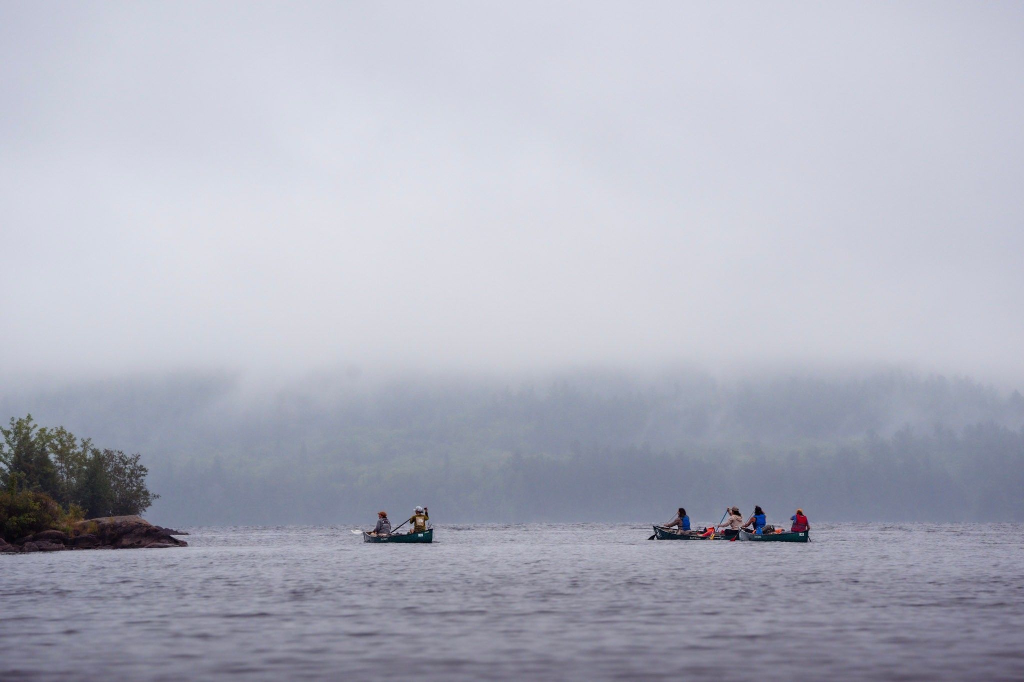 Two canoes paddle across a misty lake surrounded by fog-covered shoreline in the early morning.