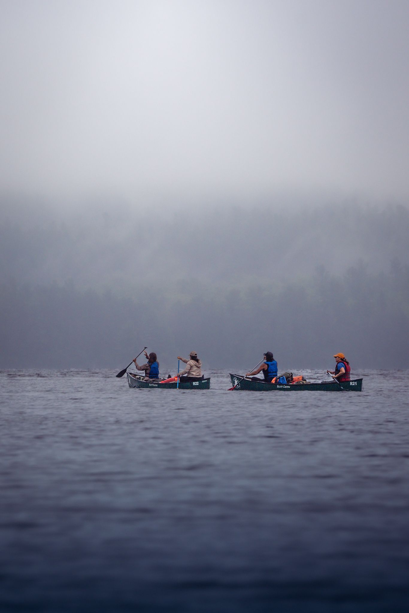 Two canoes with people paddling across a misty lake on a foggy morning with dark waters and forest in background.