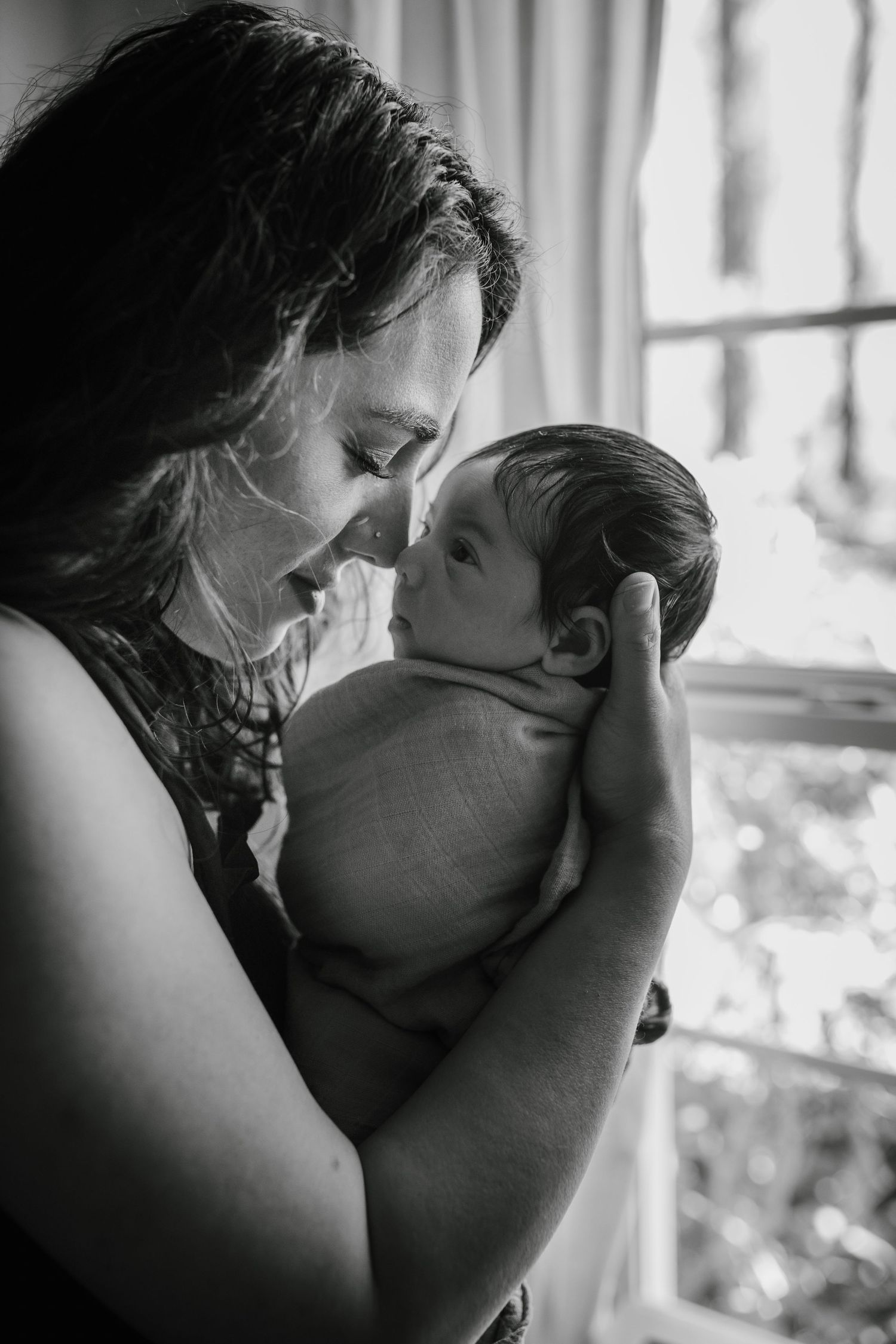 A tender black and white portrait of a parent and baby sharing an intimate moment by a window.