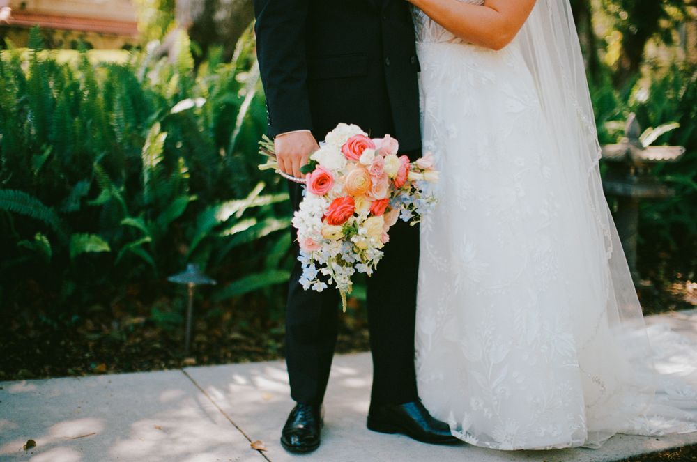 A close-up photo shows a bridal bouquet of coral roses and white flowers between two people in wedding attire.