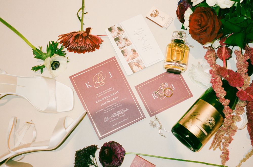 Wedding invitation cards in rose gold with floral decorations, perfume bottle and scattered flowers on white surface.