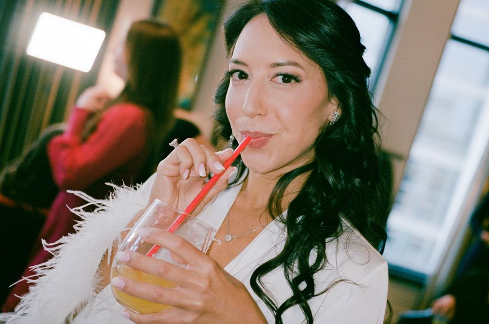 Woman in white furry jacket sips from a straw while holding a beverage at an indoor venue.