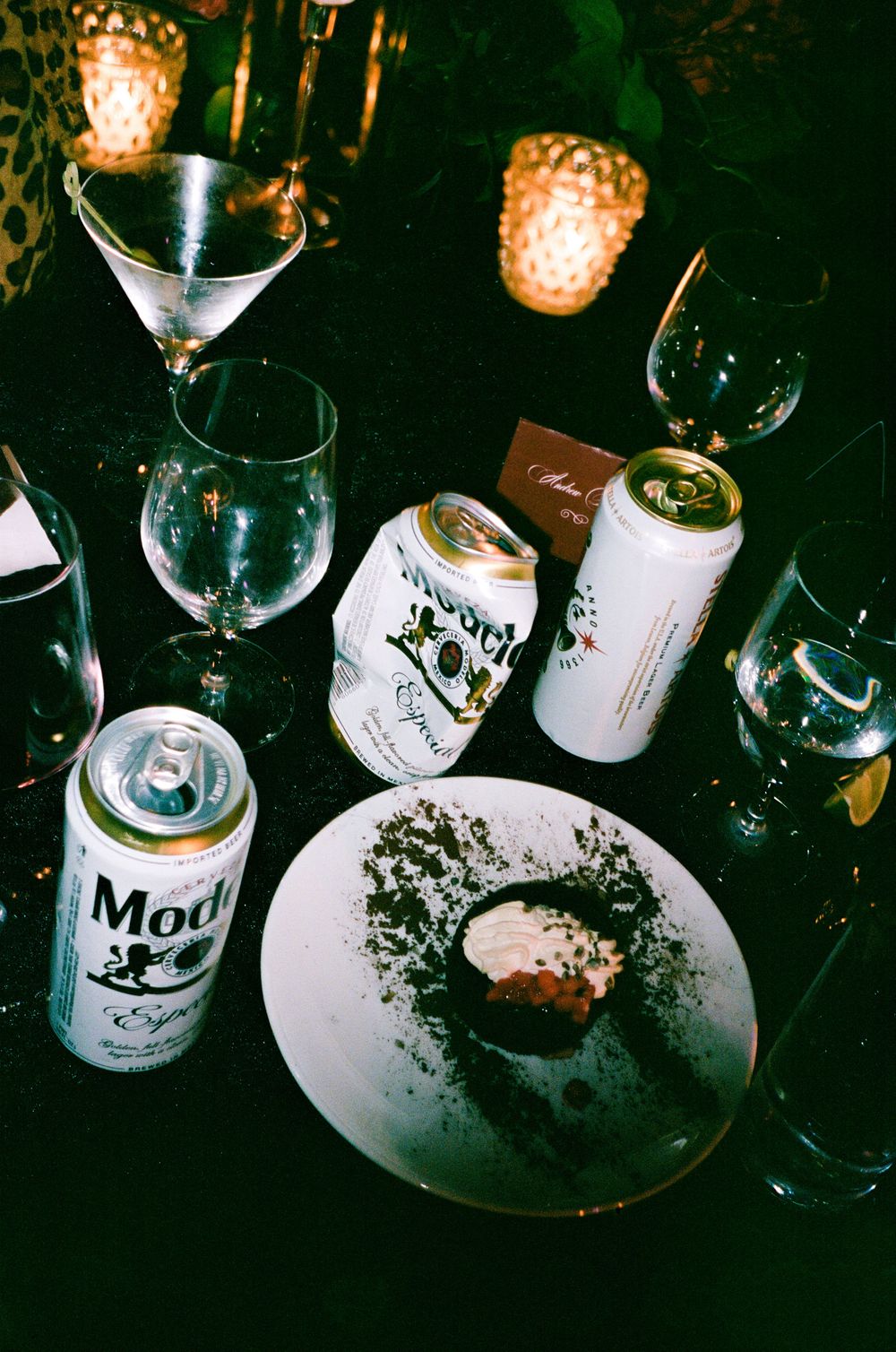 Dark moody bar setting with wine glasses and white bottles arranged on a reflective surface with ambient lighting.