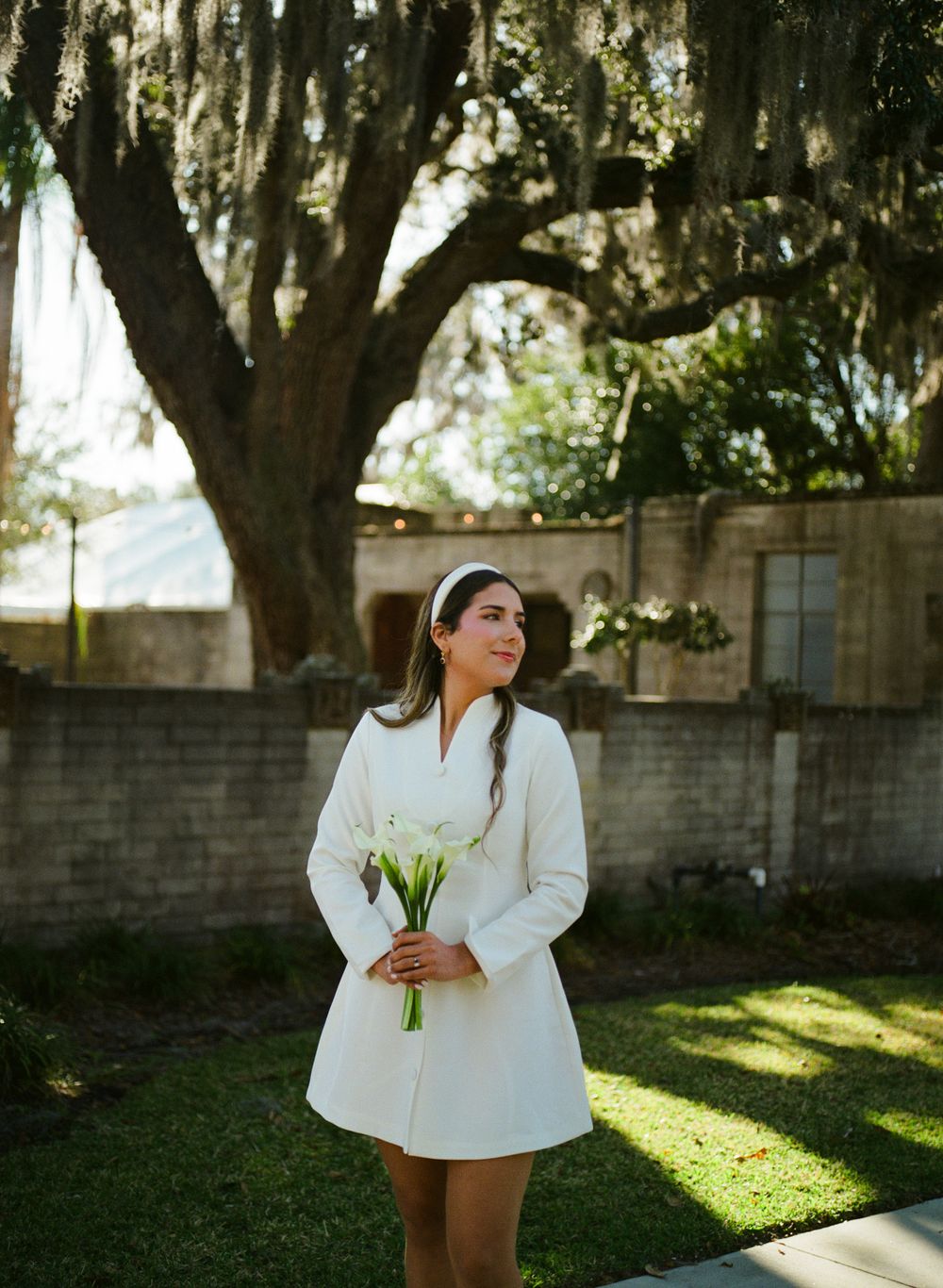 A person in a white blazer stands in a sunlit garden setting with Spanish moss hanging from trees in the background.