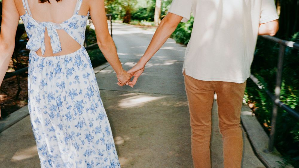 A couple in coordinating blue and white outfits walks hand in hand down a sunny garden path.
