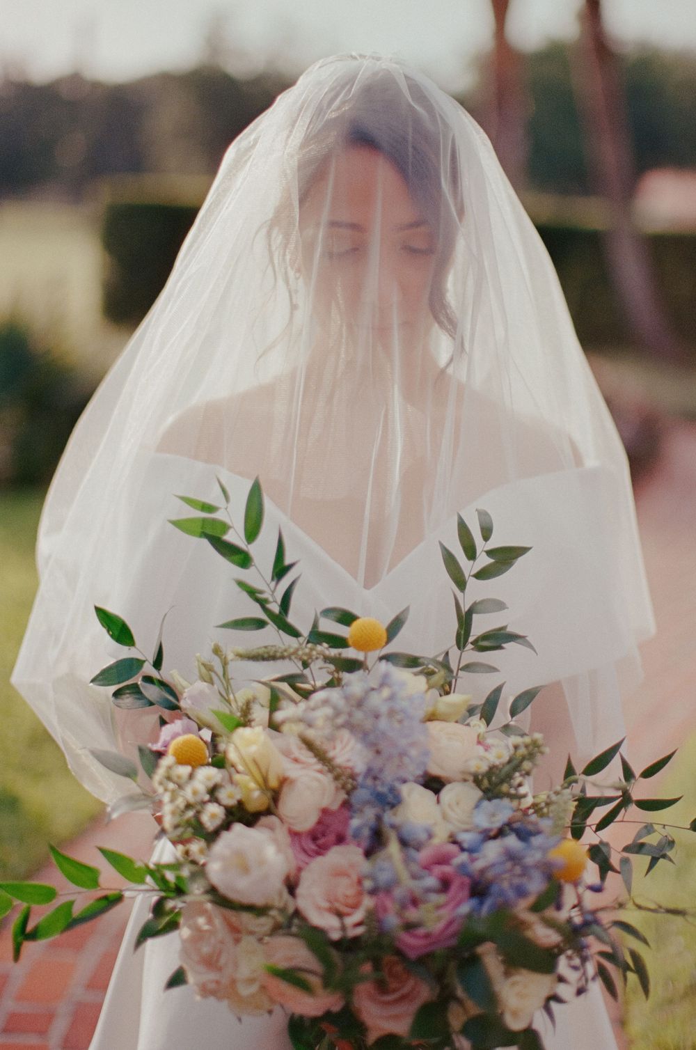 A bride in a white veil holds a stunning bouquet of pink, purple and white flowers with green foliage in an outdoor setting.