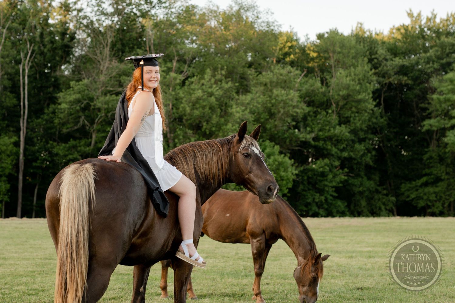 A graduate on horseback watches as another horse grazes in a green field at sunset.