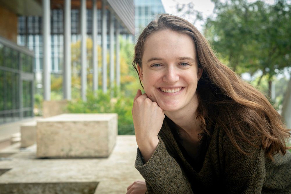 A cheerful person with long brown hair smiles while resting their chin on their hand outside a modern building.