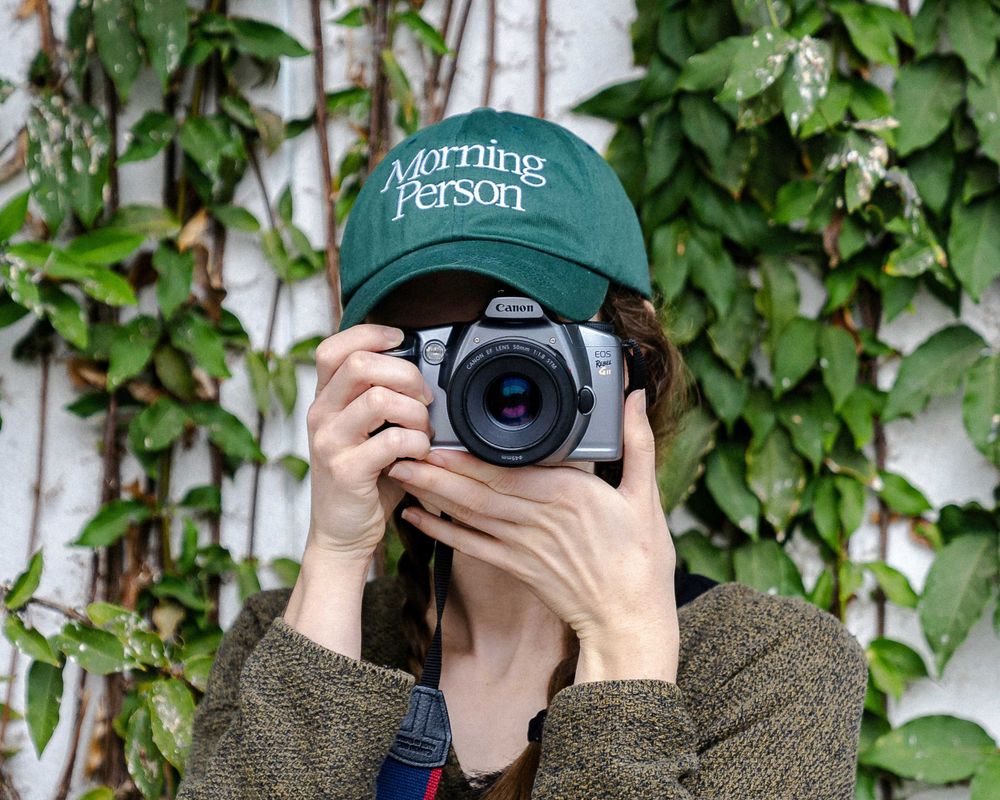 Photographer wearing green baseball cap holding camera in front of ivy-covered wall.