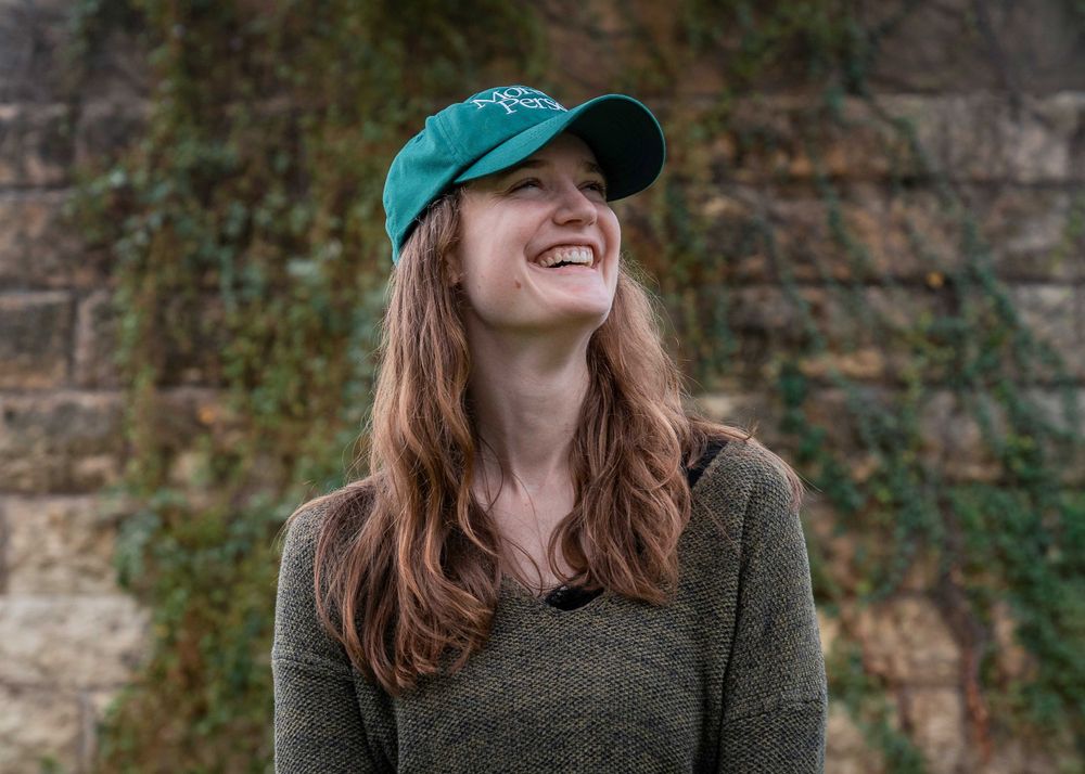 Person wearing a green baseball cap and green sweater laughs joyfully outdoors against a stone wall backdrop.
