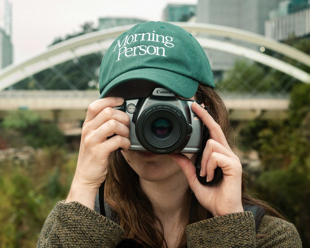 A photographer wearing a green baseball cap takes a photo with a DSLR film camera outdoors in downtown Austin, TX.