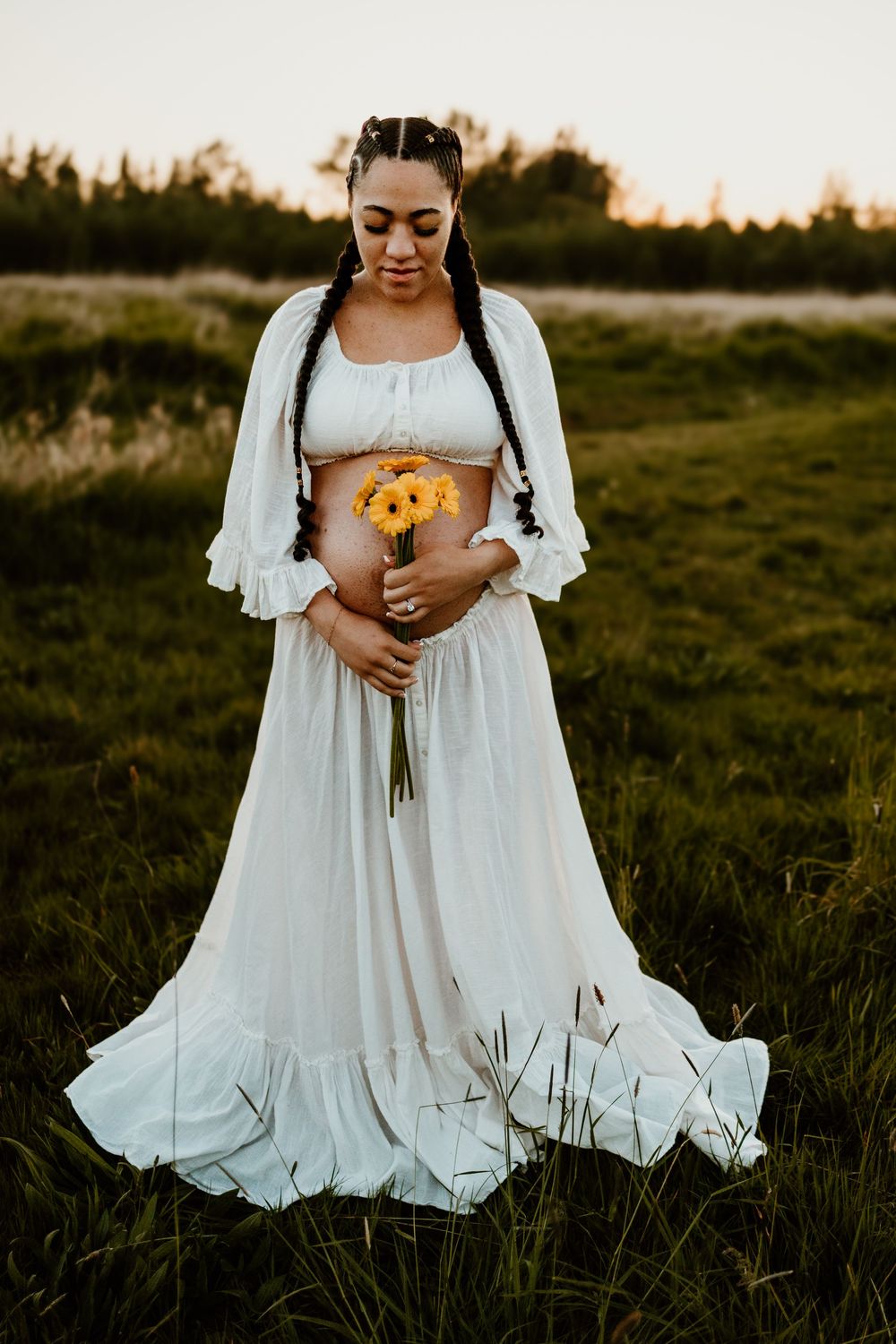 A person in a flowing white maxi dress holds sunflowers while standing in a grassy field at sunset.