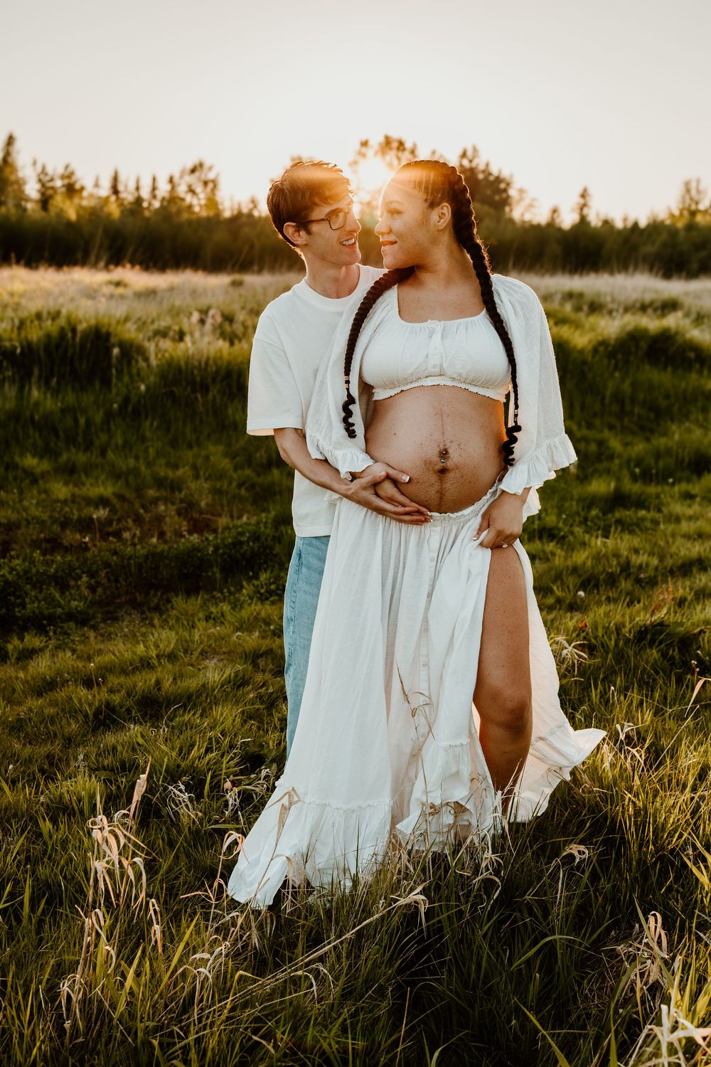 A couple shares an intimate moment at sunset in a grassy field during a maternity photoshoot.