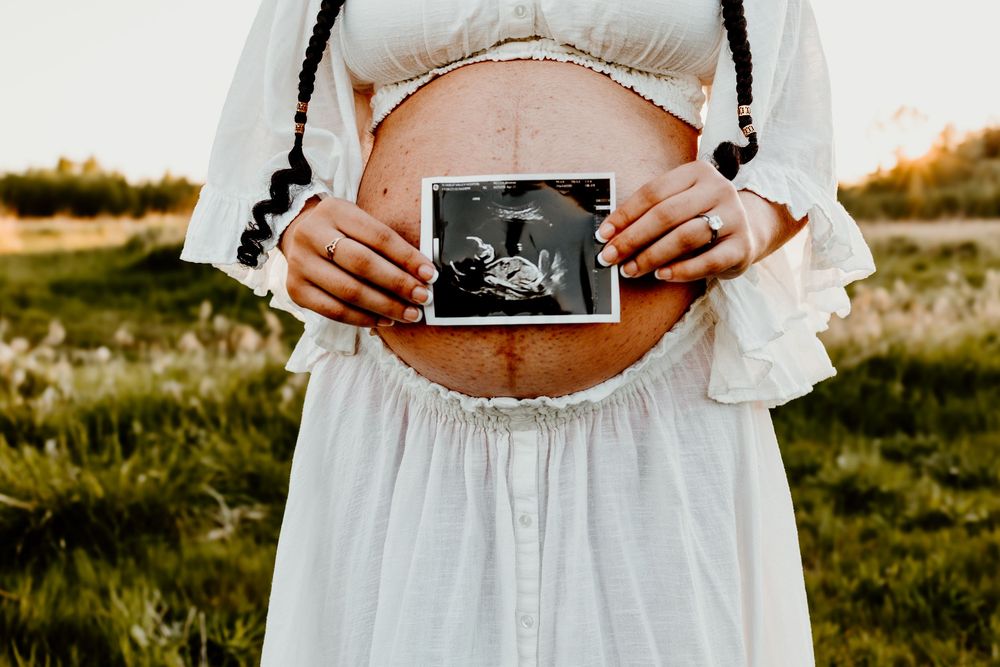 Person in white dress holds ultrasound photo against pregnant belly while standing in sunlit grassy field.
