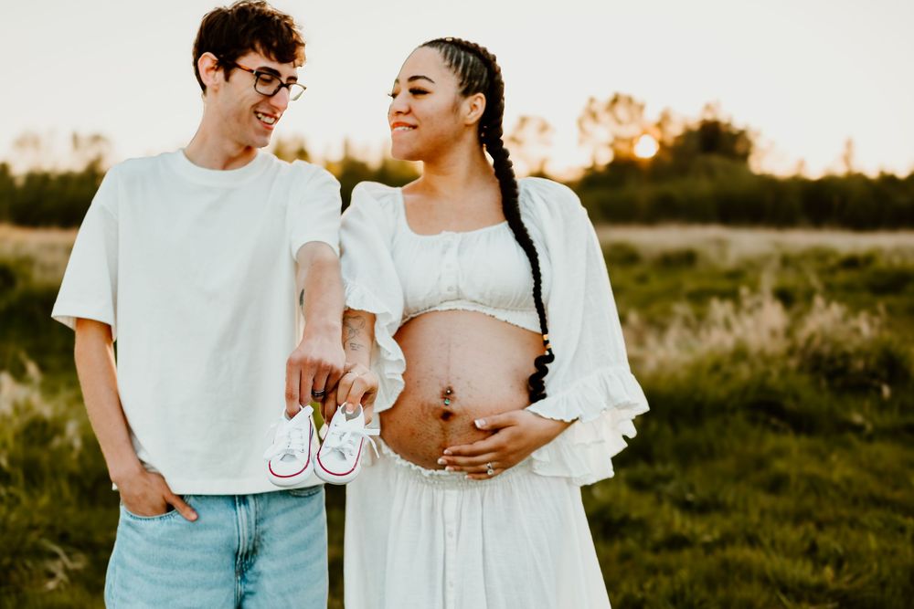 A couple stands in a grassy field at sunset holding tiny baby shoes while displaying a pregnant belly.