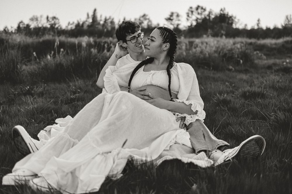 Black and white photo of two people in vintage style white dresses lounging romantically in a grass field at dusk.