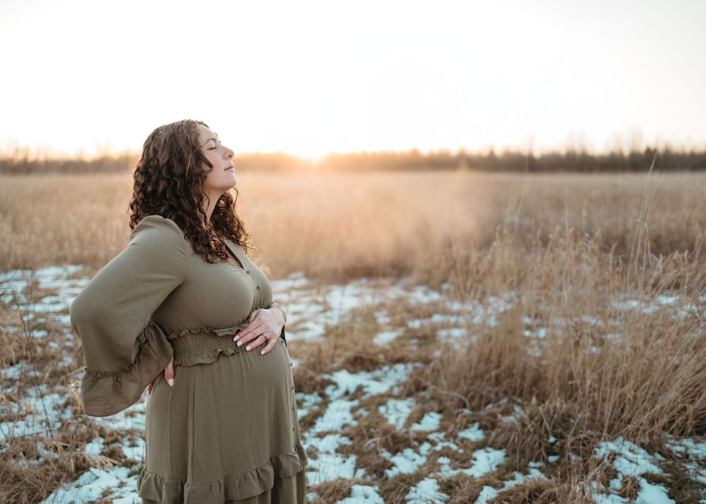 Silhouette of pregnant woman in beige dress standing in snowy field at golden sunset with tall grass in background.