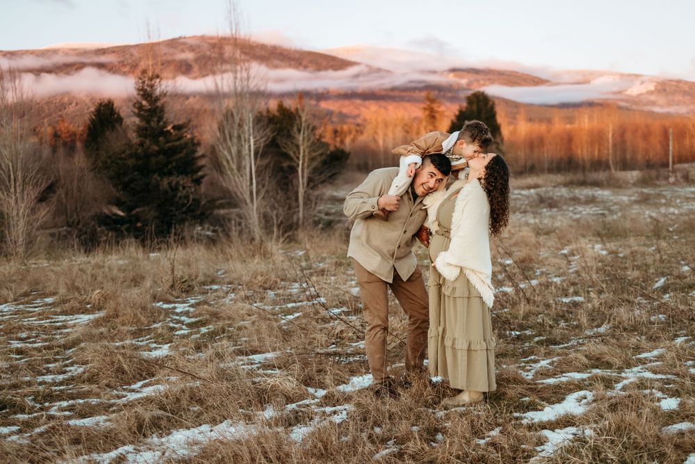 A couple embraces in a rustic field at sunset with mountains and bare trees in the background.