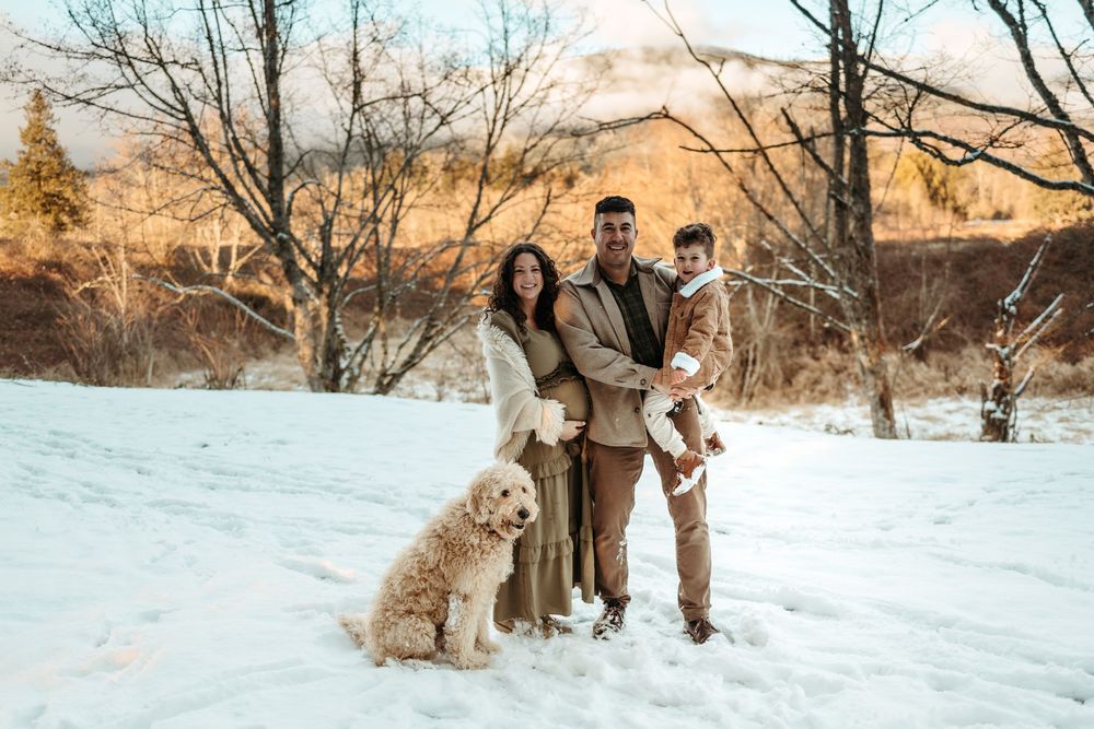 Group stands together on snowy ground with their golden doodle dog during a winter photo session at sunset.