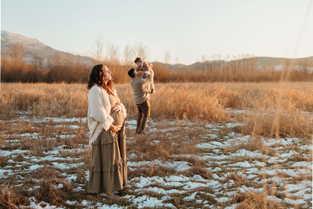 People stand in a golden field with patchy snow at sunset, surrounded by mountains and winter prairie grass.
