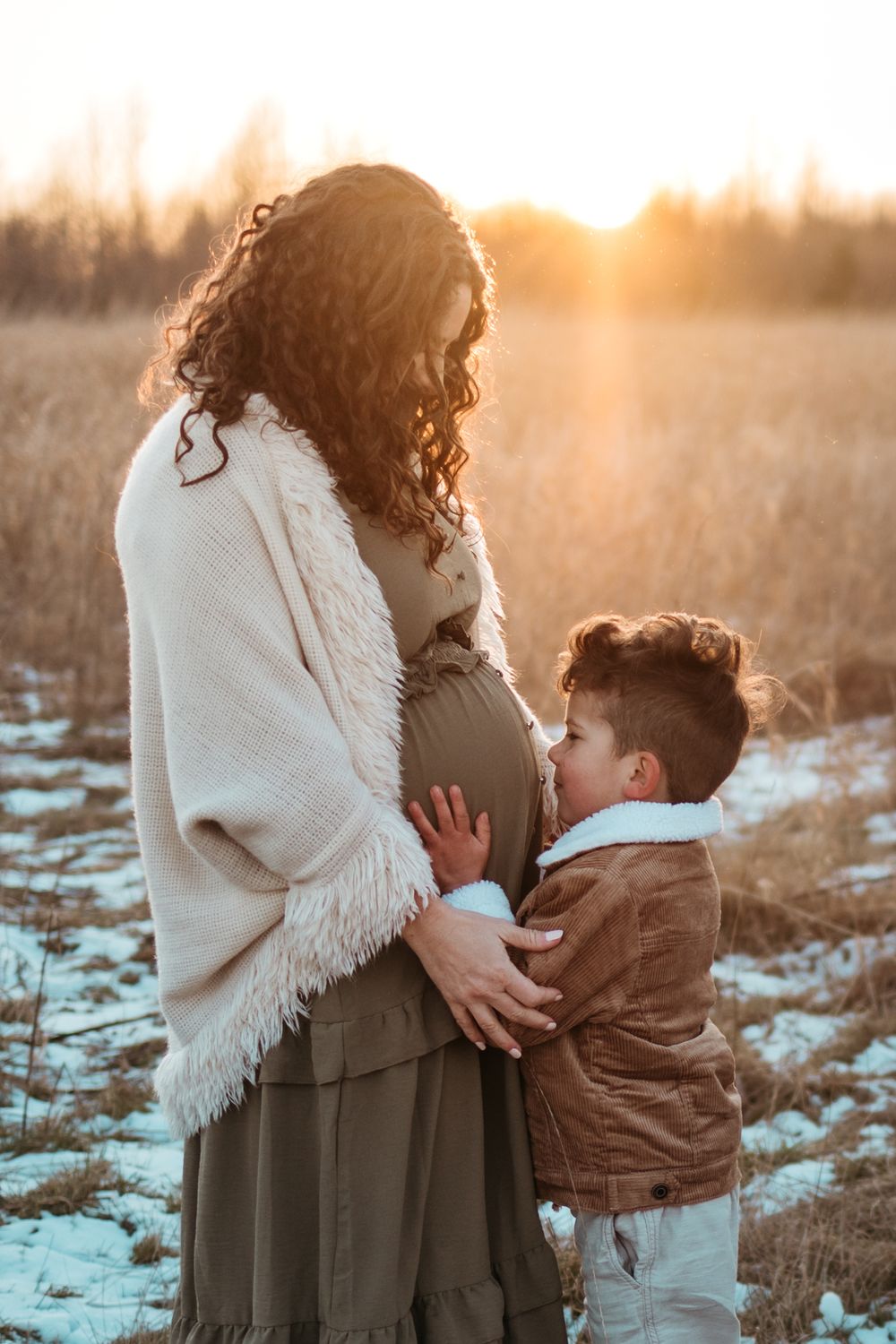 A tender moment between a pregnant woman in a white shawl and a young child at sunset in a snowy field.