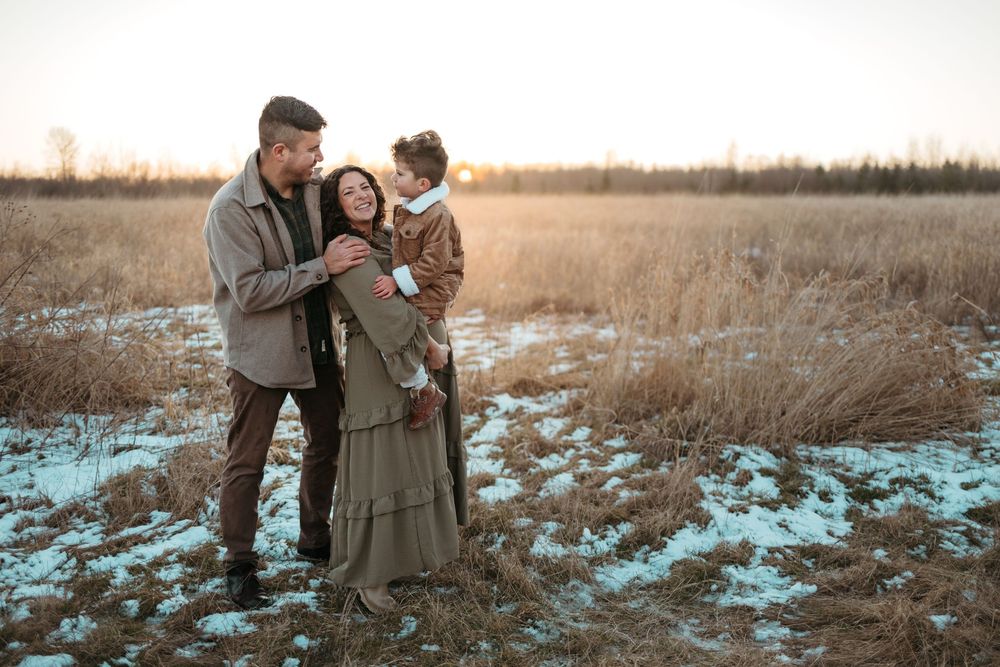 Family standing together in winter field with snow patches and tall grass at golden sunset.