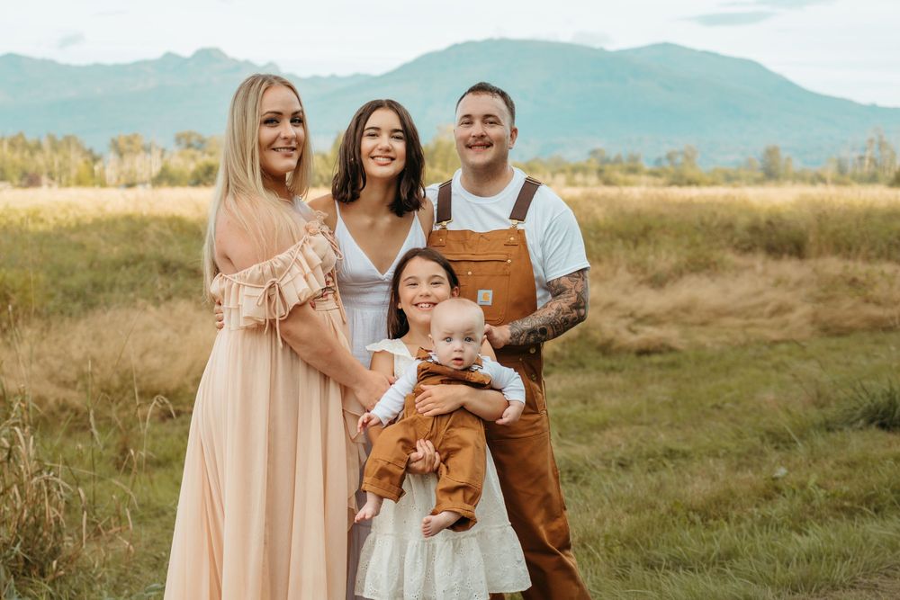 A family in neutral tones poses together in a golden field during sunset with mountains in the background.