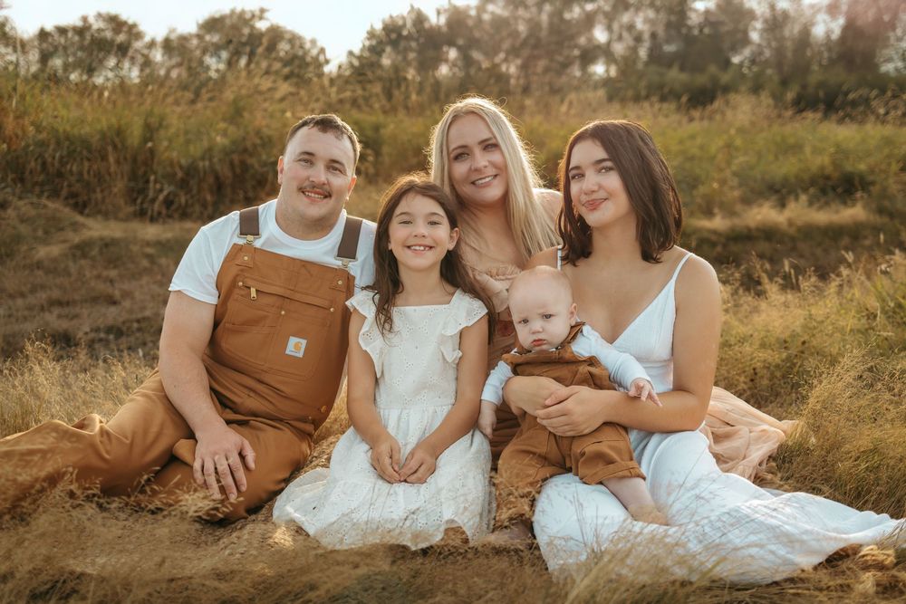 Family sitting together in golden field at sunset wearing coordinated white and brown outfits.