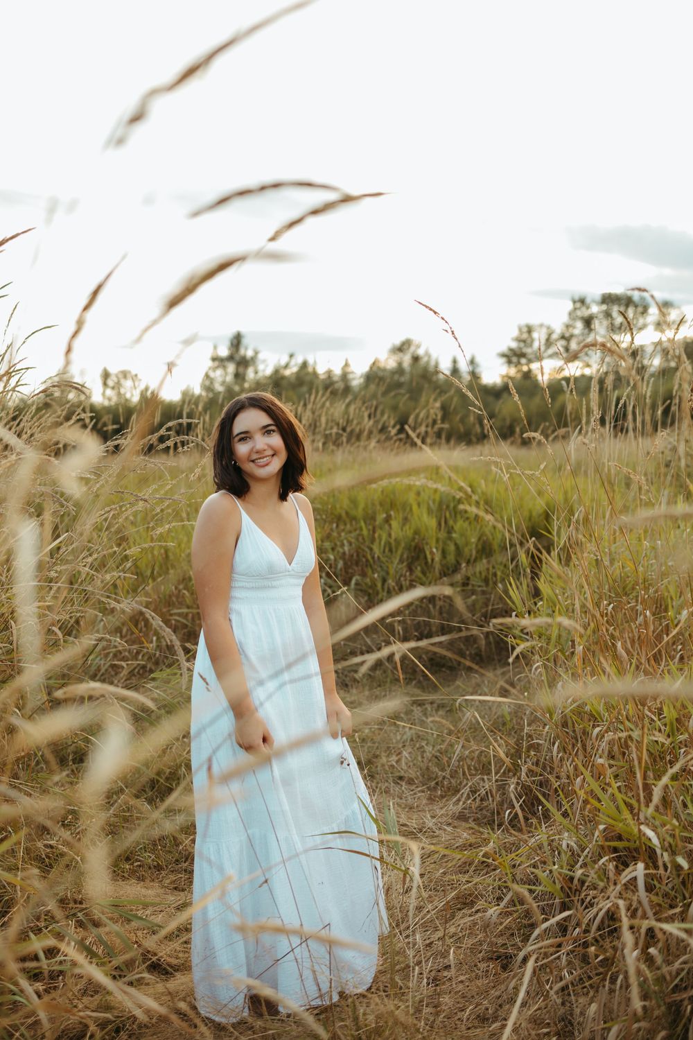 A figure in a light blue sundress stands gracefully among tall beach grass at sunset, creating a dreamy coastal scene.