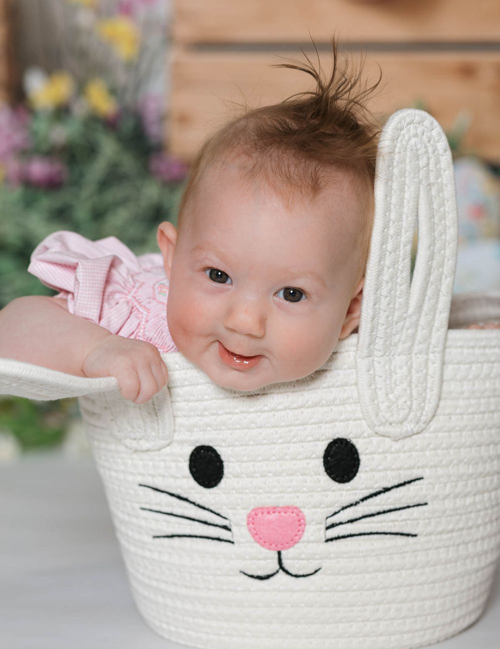 Smiling baby in pink resting inside a white knitted bunny-shaped basket with flowers in background.