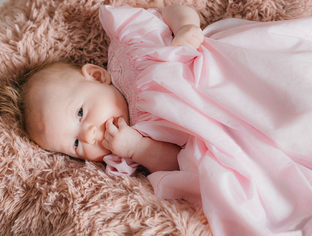 Small infant wearing pink dress lies comfortably on soft beige plush blanket.
