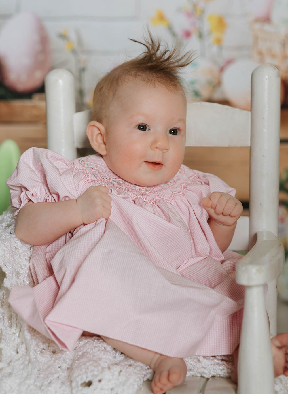 A baby in a pink smocked dress sits in a white wooden chair against a soft pastel background.