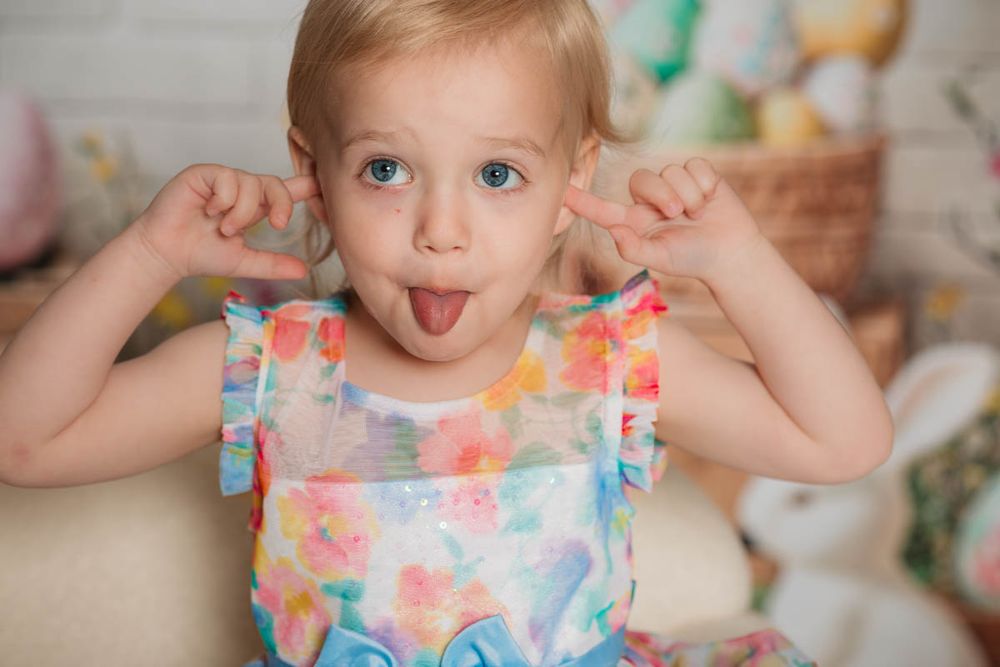 A playful toddler wearing a pastel tie-dye dress sticks out their tongue while raising both hands by their ears.