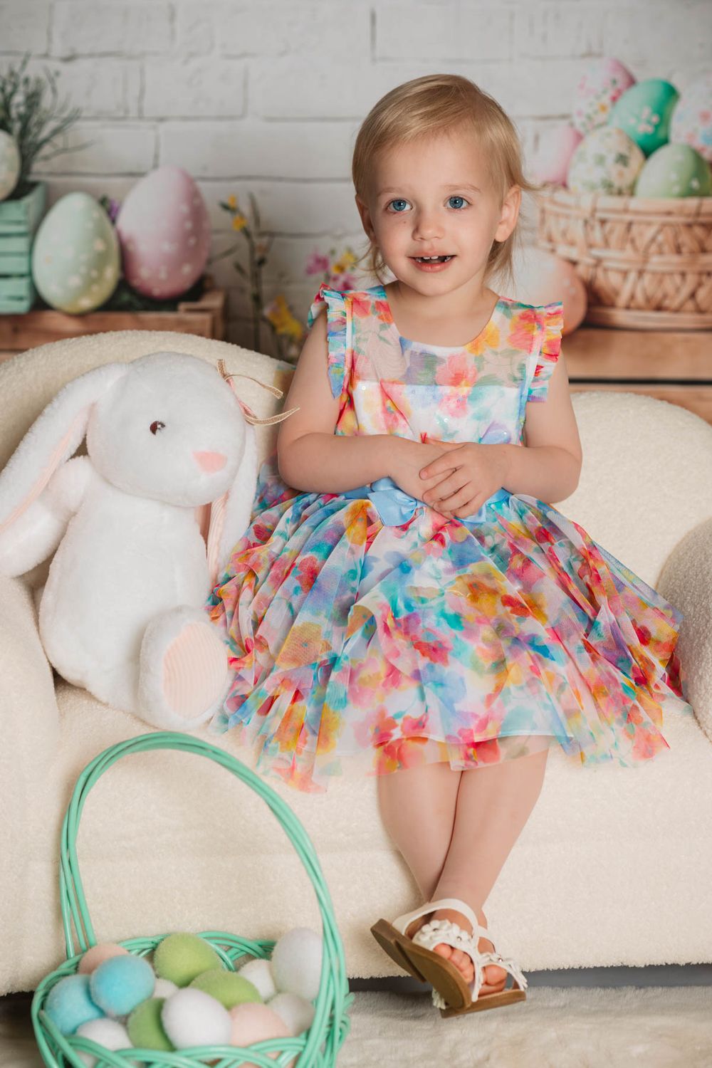 Easter holiday photo with colorful floral dress, plush bunny, pastel eggs in basket and white brick backdrop.